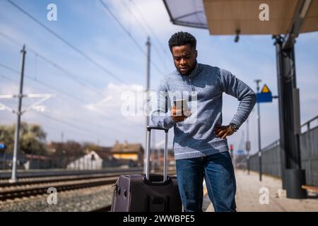 Uomo scioccato con una valigia e un telefono cellulare in piedi su una stazione ferroviaria. Foto Stock