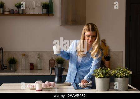 Donna con caffè in una cucina moderna. La ragazza tiene in mano una tazza di caffè e pulisce la superficie del piano cottura Foto Stock