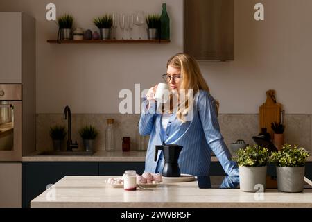 Donna che sorseggia un caffè in cucina alla luce del sole. Routine mattutina Foto Stock