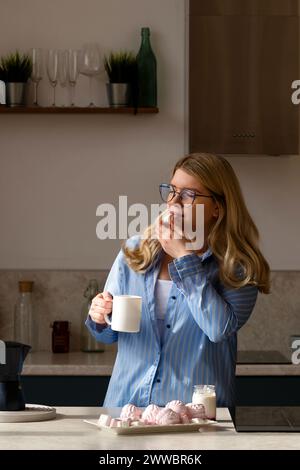 Donna che assaggia il caffè, cucina tranquilla. Una ragazza con camicia a righe blu ama i marshmallow sorseggiando un caffè mattutino Foto Stock