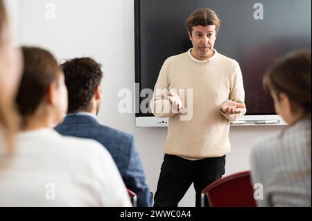 Uomo di mezza età in piedi vicino a una lavagna interattiva e in comunicazione con studenti adulti durante una lezione di lingua straniera Foto Stock