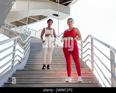Due donne in abbigliamento fitness con colori diversi che guardano la fotocamera mentre sono in piedi su una scala. Donna di grandi dimensioni con la sua amica del fitness che posa Foto Stock