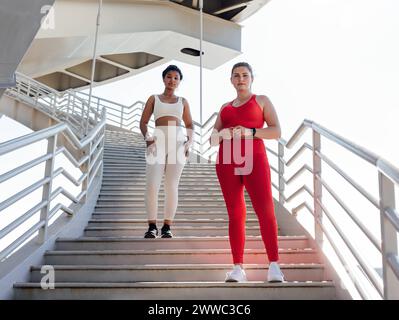Due donne in abbigliamento fitness con colori diversi che guardano la fotocamera mentre sono in piedi su una scala. Donna di grandi dimensioni con la sua amica del fitness che posa Foto Stock