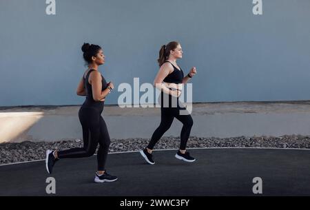 Due donne con diversi tipi di corpo che corrono all'aperto. Vista laterale di due giovani donne che fanno jogging su una parete grigia. Due donne con diversi tipi di corpo runni Foto Stock