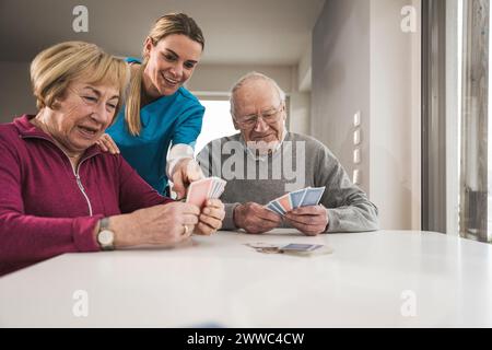 Felice coppia senior che gioca a carte con l'assistente domestico a tavola Foto Stock