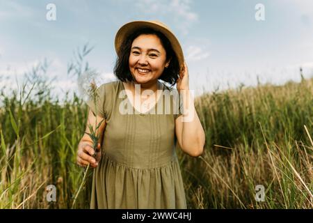 Donna matura felice che indossa un cappello e tiene il dente di leone sul campo Foto Stock