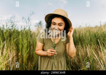 Donna giocosa che indossa un cappello e tiene il dente di leone vicino al viso in campo Foto Stock
