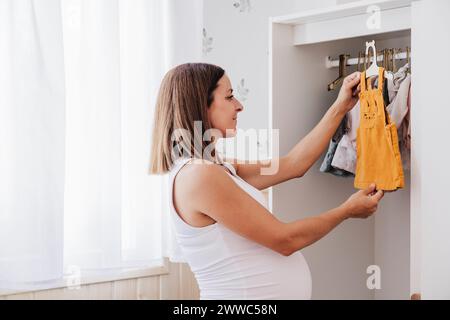 Aspettarsi una donna incinta che sistemi vestiti per bambini nell'armadio a casa Foto Stock