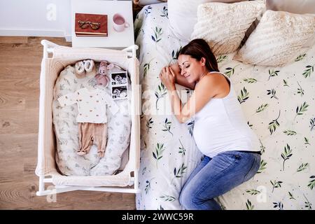 Donna incinta che dorme sul letto vicino alla culla di casa Foto Stock