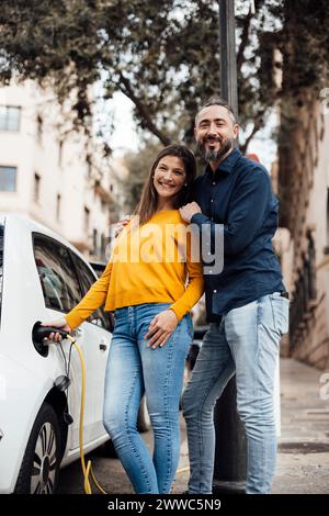 Donna sorridente che carica l'auto elettrica e sta con l'uomo Foto Stock