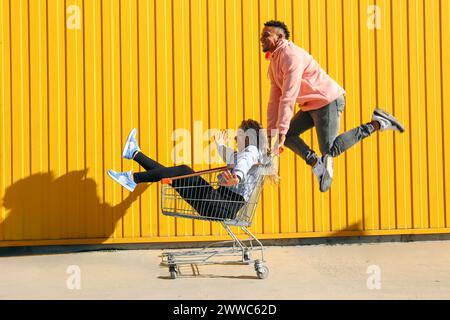 Donna spensierata seduta nel carrello vicino a un uomo che salta di fronte a un muro a strisce gialle Foto Stock