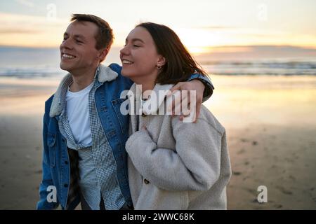 Fidanzato sorridente e fidanzata che camminano con le braccia intorno alla spiaggia dell'oceano Foto Stock