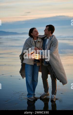 Una giovane coppia sorridente con una lanterna che cammina in scialle sulla spiaggia Foto Stock