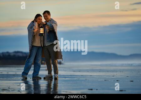 Coppia sorridente con lanterna in piedi sulla spiaggia al tramonto Foto Stock