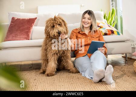 Bella donna felice che tiene in mano un tablet PC e accarezza un cane a casa Foto Stock