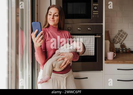 Donna sorridente che porta la bambina e usa lo smartphone in cucina Foto Stock