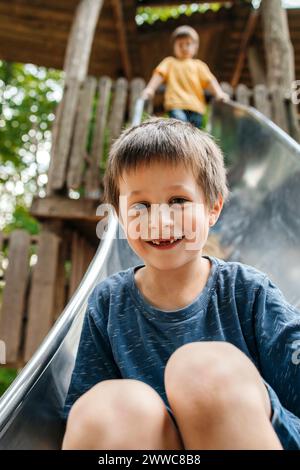 Il ragazzo felice scivola giù sullo scivolo con il fratello sullo sfondo al parco giochi Foto Stock