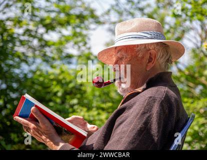 Uomo anziano che fuma pipa e legge un libro in giardino Foto Stock