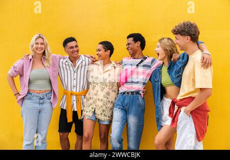 Amici spensierati che indossano abiti colorati in piedi davanti alla parete gialla con le braccia intorno Foto Stock