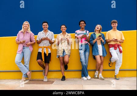 Gruppo di amici appoggiati contro il muro giallo utilizzando il proprio smartphone Foto Stock