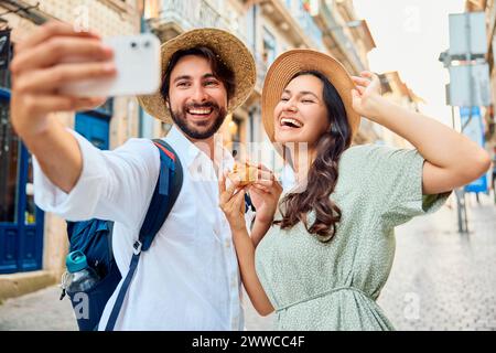 Una giovane coppia sorridente che indossa un cappello e scatta selfie con il tradizionale dolce pastel de nata tramite smartphone Foto Stock