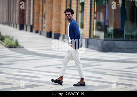 Un bell'uomo afroamericano alla moda cammina lungo la strada con eleganti pantaloni bianchi, blazer blu e mocassini. Tendenza alla moda per lo Street style Foto Stock