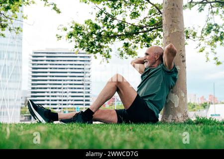 Anziano in pensione appoggiato sul tronco dell'albero con le mani dietro al parco Foto Stock