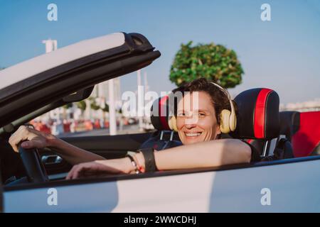 Donna sorridente che indossa le cuffie wireless in un'auto convertibile nelle giornate di sole Foto Stock