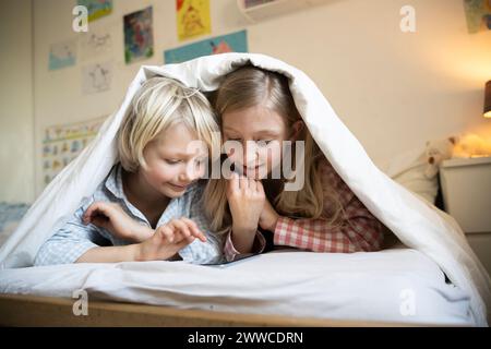 Fratello e sorella guardano lo smartphone sdraiati sotto la coperta sul letto a casa Foto Stock