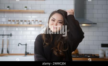 Felice donna asiatica seduto in cucina a guardare la macchina fotografica e sorridere. Donna accogliente che si rilassa in casa Foto Stock