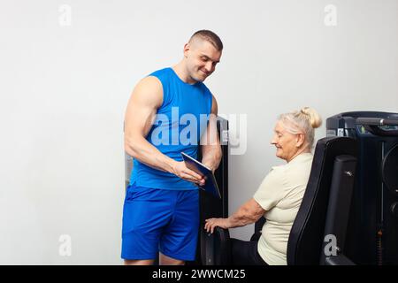 Un giovane e affascinante allenatore mostra alla donna più anziana come eseguire esercizi, immagini in tablet. Tecnologie attuali in palestra. Il coach registra il Foto Stock