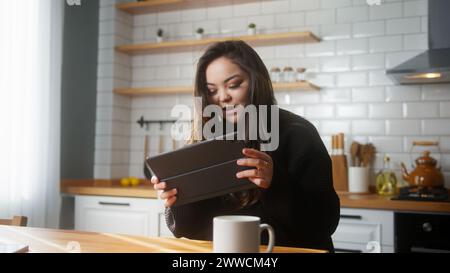 Ragazza asiatica che gioca a un videogioco su un tablet mentre è seduta in cucina a casa. Donna eccitata e concentrata, coinvolta in un gioco interessante Foto Stock