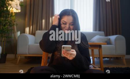 Donna asiatica sorridente seduta sul pavimento a scrivere un messaggio di testo tramite smartphone. Donna che si rilassa nel soggiorno di casa, comunica con gli amici Foto Stock