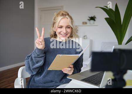 L'immagine di una ragazza sorridente registra video di se stessa su una fotocamera digitale, mostra un segno di pace, si siede davanti al laptop, tiene il notebook. Foto Stock