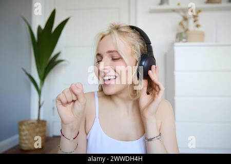 Ritratto di una donna felice, ascolta la musica con le cuffie wireless, tiene in mano il microfono invisibile, canta nel microfono, sorride, goditi una buona qualità audio Foto Stock