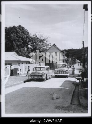 Descrizione: Honduras britannico. [Scena di strada a Benque Viejo, del Carmen Cayo District. L'immagine mostra due auto. Uno sta guidando verso la telecamera mentre l'altro è parcheggiato con una famiglia che cammina verso di essa. Un cane sta sdraiando sulla strada]. Fotografia n. R 34997 Official British Honduras Fotografia compilata dall'Ufficio centrale delle informazioni. Copyright Crown riservato. Luogo: Honduras britannico Data: 1965 Apr belize, caraibi, caribbeanthrough alens Foto Stock