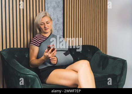 una bionda con un bendaggio su una mano e tenendo un telefono in un'altra mentre si siede sul divano, assistenza sanitaria domiciliare. Foto di alta qualità Foto Stock