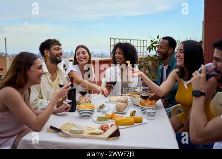 Gruppo ridendo di diversi giovani amici gustando il pranzo insieme all'aperto celebrando la festa sul tetto. Foto Stock