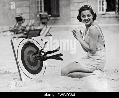 Hollywood, Los Angeles, California, c. 1927 Una Martha Sleeper sorridente colpisce i bullseyes del bersaglio con freccette sulla spiaggia. Foto Stock