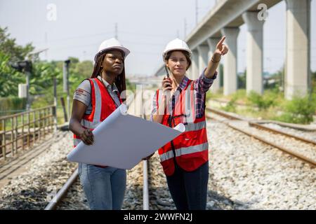 Due costruttori o ingegneri con casco di sicurezza e giubbotto ad alta visibilità che esaminano i lavori da eseguire o i progressi su un progetto di binario ferroviario durante l'holdin Foto Stock