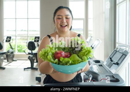 Una signora di grandi dimensioni in piedi su un tapis roulant, mantenendo la sua salute con una ciotola di frutta e verdura fresca. Foto Stock