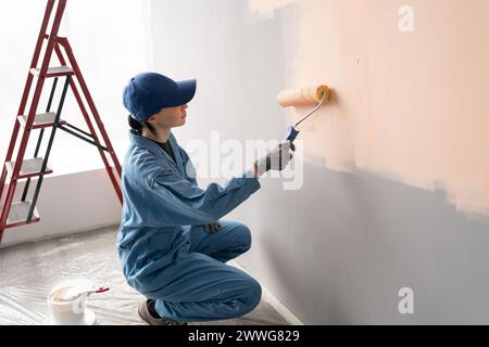 vista dall'alto di una pittrice in tuta con cappello e guanti che dipinge una parete con rullo per pittura, scala e secchi sullo sfondo Foto Stock