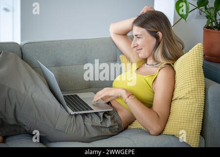 Lavoratrice freelance di mezza età rilassata online che lavora su un notebook, sdraiata sul divano di casa Foto Stock