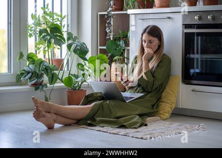 Una donna preoccupata distratta dal portatile sullo smartphone riceve cattive notizie in un messaggio seduto sul pavimento di casa. Foto Stock