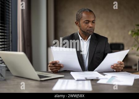 Un uomo d'affari serio e fiducioso finanziatore all'interno dell'ufficio sul posto di lavoro che controlla documenti, rapporti e contratti. Uomo anziano in tuta aziendale che lavora su documenti, contabile con conti. Foto Stock