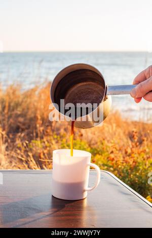 l'uomo versa il caffè appena estratto da un mestolo in alluminio in una tazza su un tavolo turistico, in riva al mare. Viaggi e turismo. Foto Stock