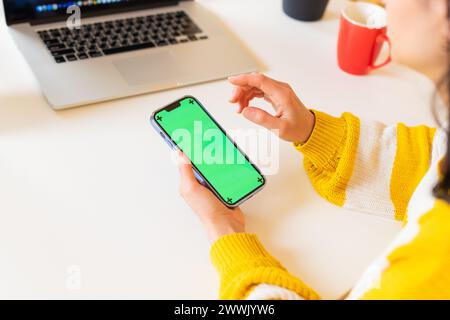 Primo piano donna irriconoscibile in un maglione che lavora utilizzando un moderno smartphone vuoto con schermo verde in un ufficio da tavolo con display cromatico Foto Stock