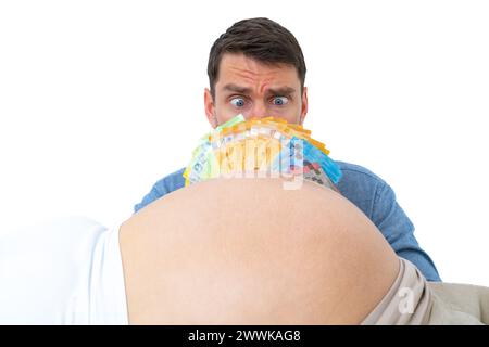 Descrizione: Il padre incinta si inchina incredibilmente davanti a una pila di banconote davanti al viso davanti al bump del bambino incinta. L'ultimo mese di pre Foto Stock