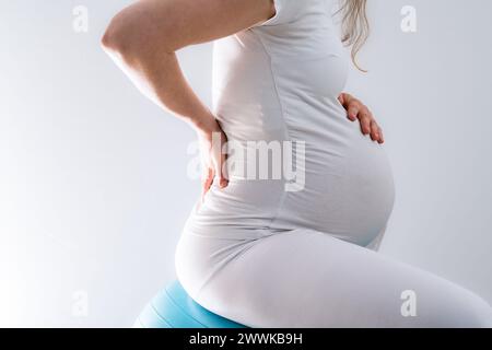 Descrizione: La madre incinta con bambole tratta il mal di schiena e con l'aiuto di una palla da palestra. Ultimo mese di gravidanza - settimana 36. Vista laterale. Bianco bac Foto Stock