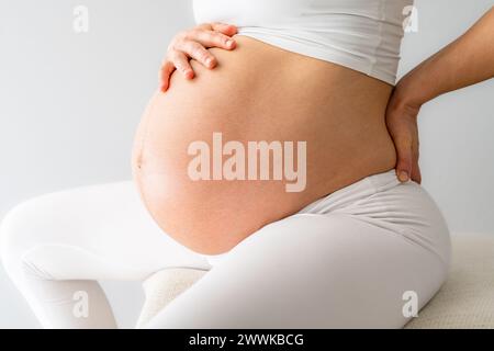 Descrizione: Parte centrale di una donna con un bambino pesantemente incinta seduta su una palla di esercizio e sensazione di mal di schiena. Vista laterale. Sfondo bianco. BR Foto Stock